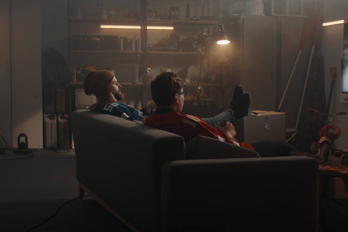 Two young men are sitting on a couch in their garage. They're looking at a game on a large, flat-screen tv.