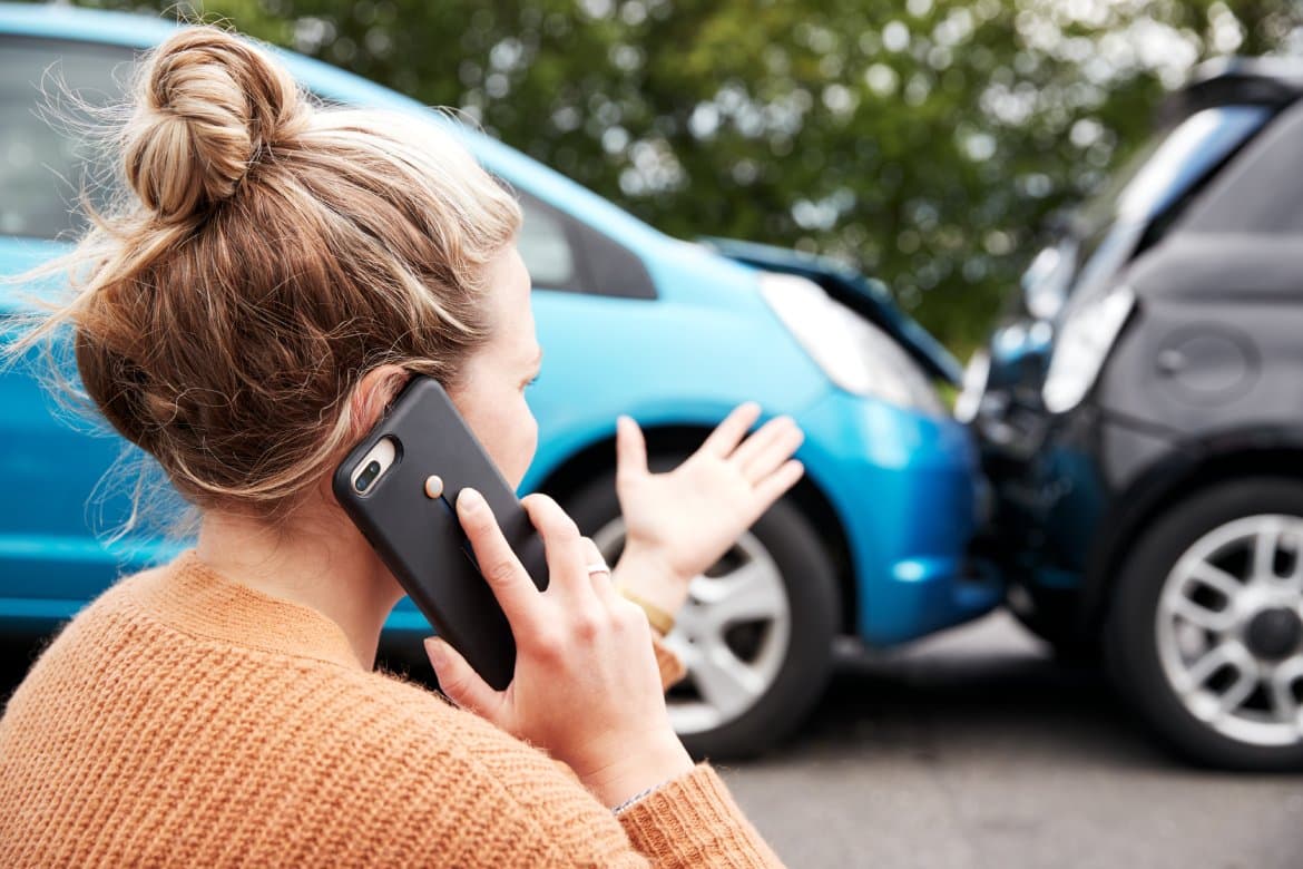 A female motorist on the phone with her auto insurance provider. She's pointing to an auto collision in front of her.