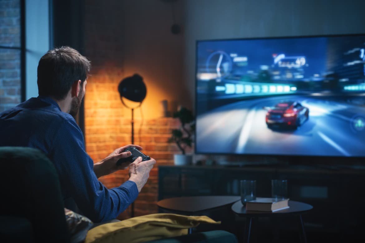 A man holds a video game controller. The tv has an animated car turning sharply. A lamp lights the corner of the room.