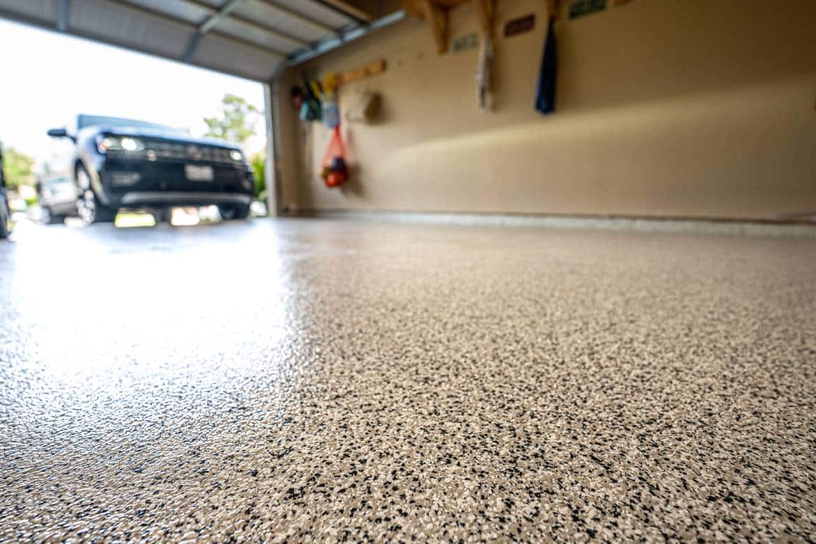A close-up view shows a shiny, brown, white, and black speckled garage floor. Blurry vehicles sit in the background.