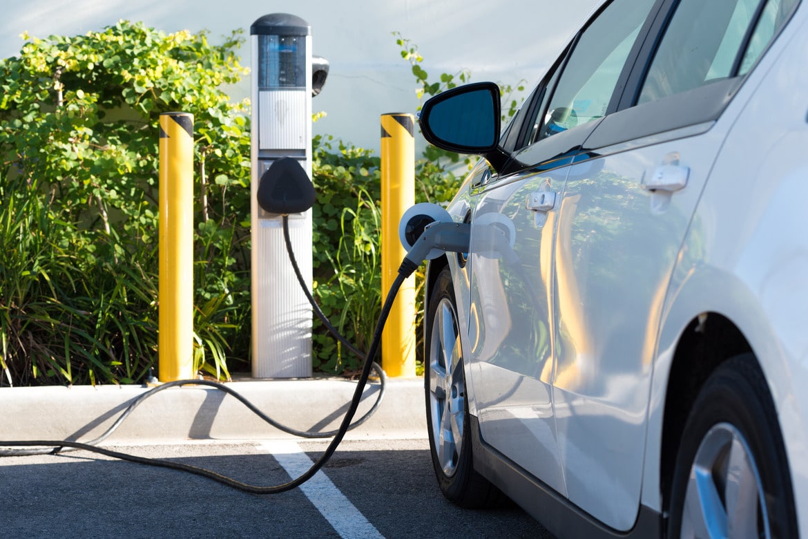 An electric vehicle parked at a public charging station on a sunny day. The ev is white and the charger is plugged in.