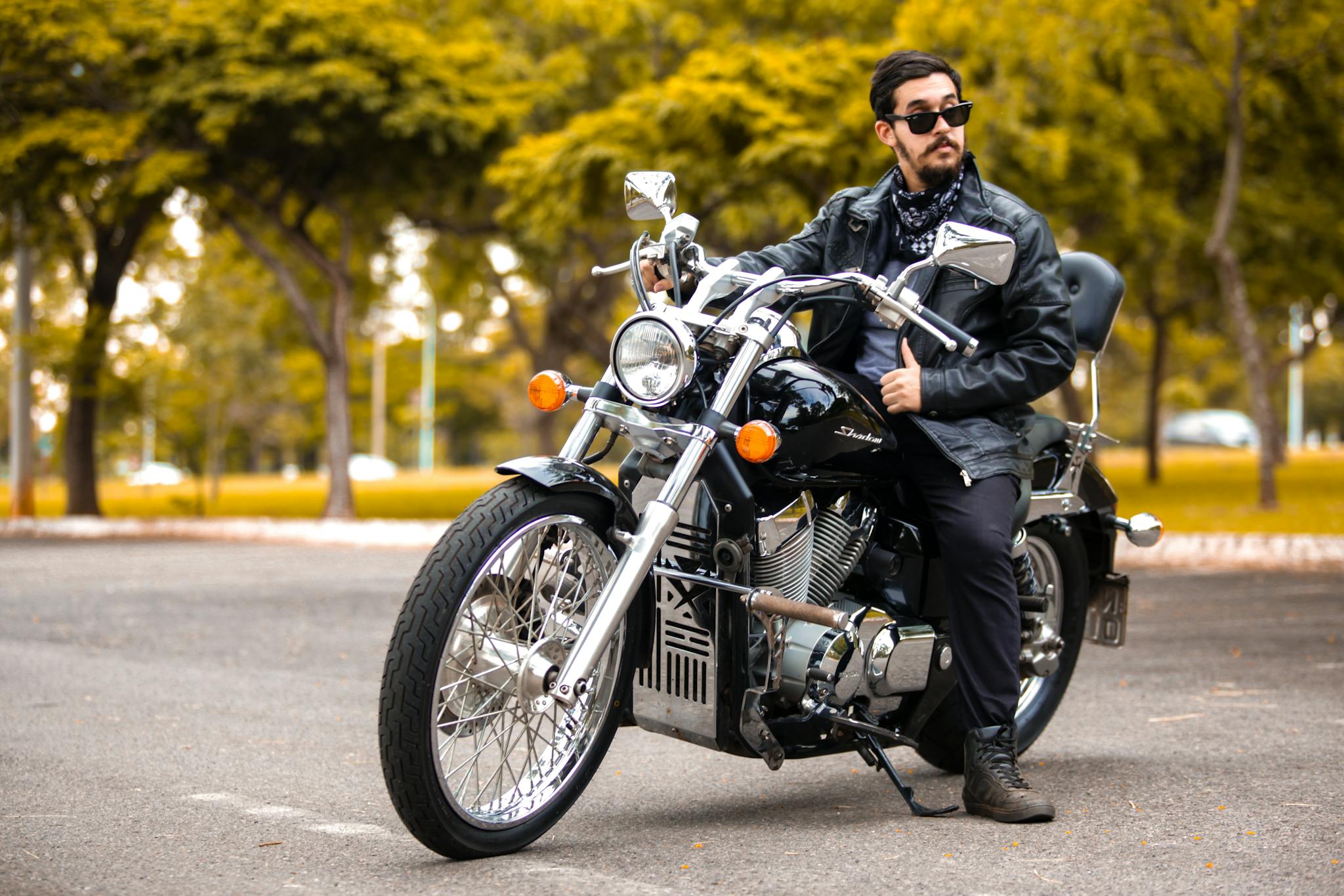 Man on vintage motorcycle in park setting, wearing sunglasses and leather jacket.