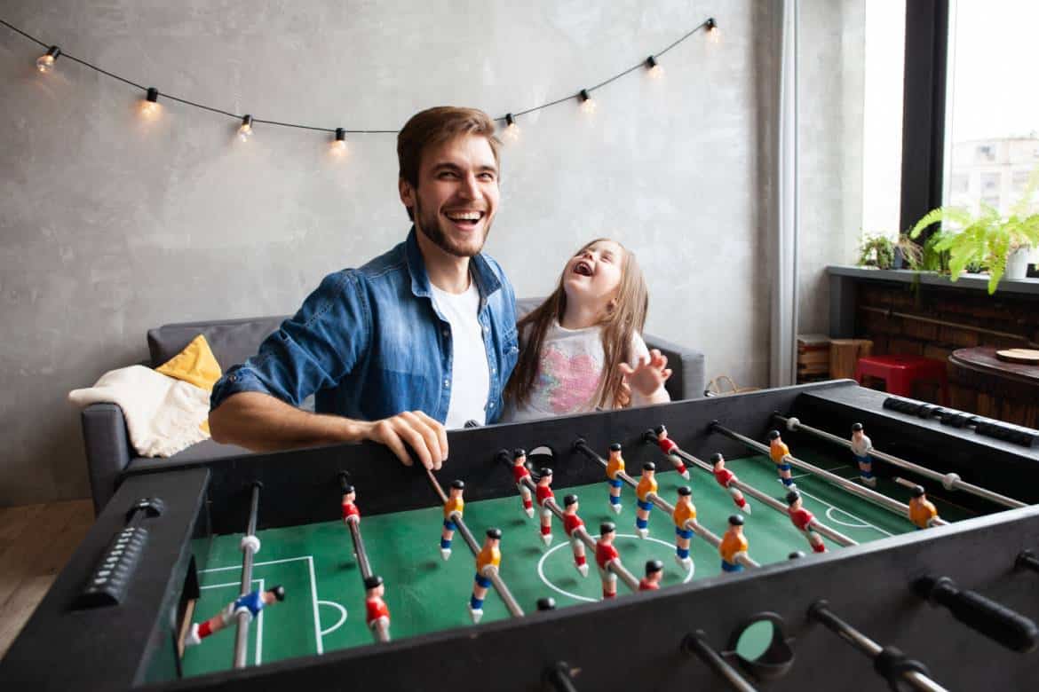 A man and a young child laugh while playing foosball. There is a couch and string lights in the background.