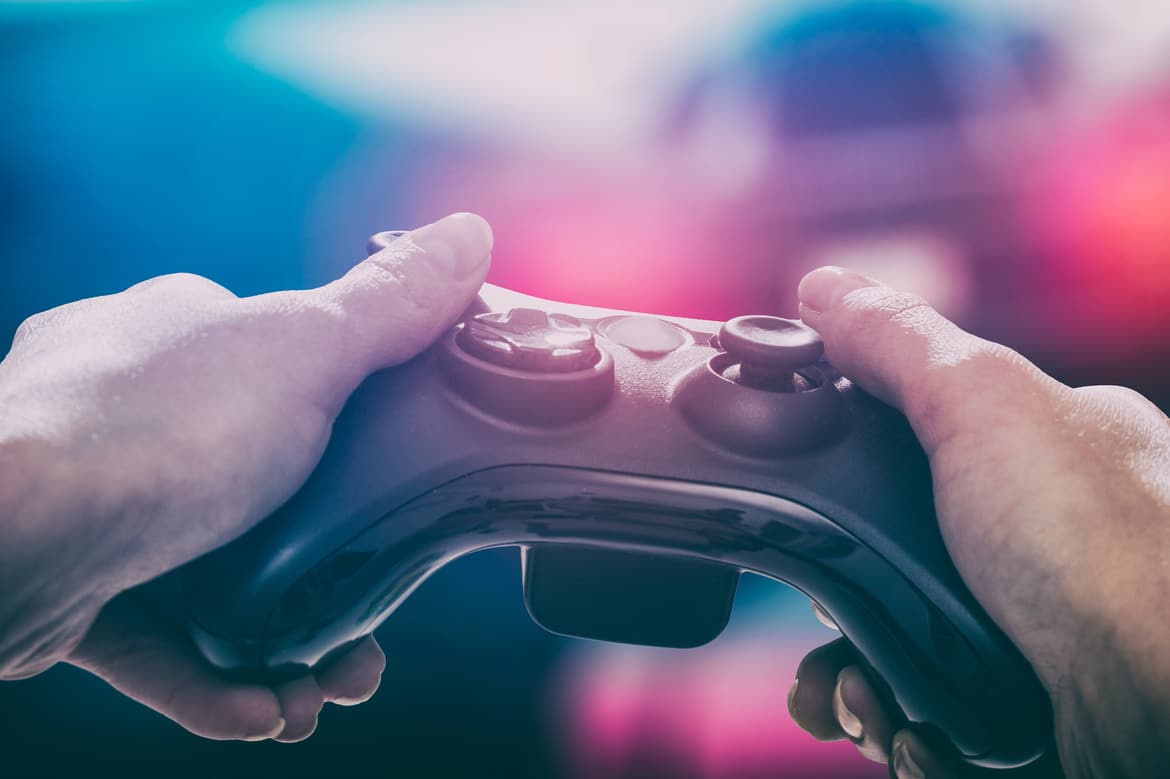 A close-up view shows a pair of hands holding a black game console controller. The background is blurry.