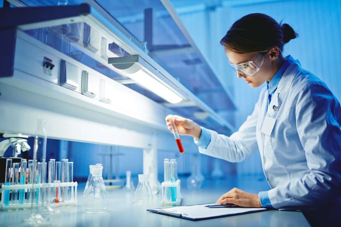 A scientist in a white lab coat and safety glasses holds up a test tube of red liquid while looking at a clipboard nearby.