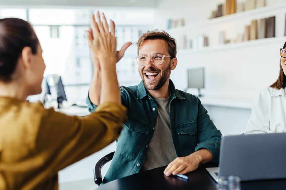 Smiling employees giving each other a high five while sitting at a table with a laptop at the workplace.