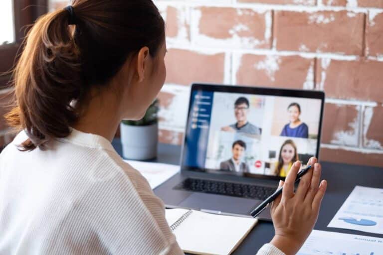A woman wearing a white sweater and holding a pen is sitting at a desk talking to four people through a small laptop.
