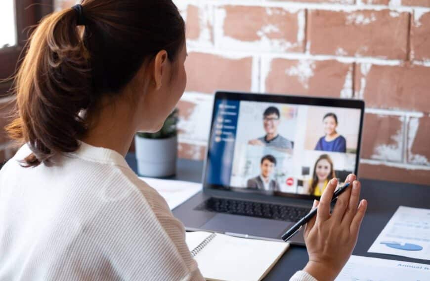 A woman wearing a white sweater and holding a pen is sitting at a desk talking to four people through a small laptop.