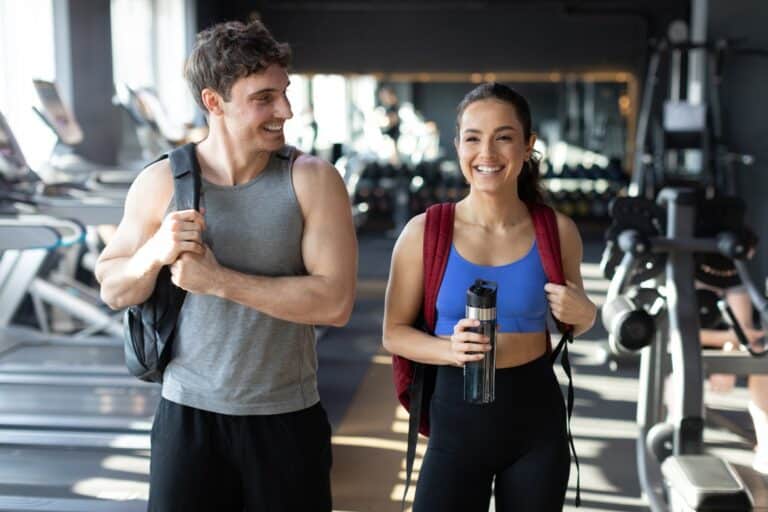 A man and a woman walk through a modern gym dressed for exercise while holding a water bottle and backpacks.