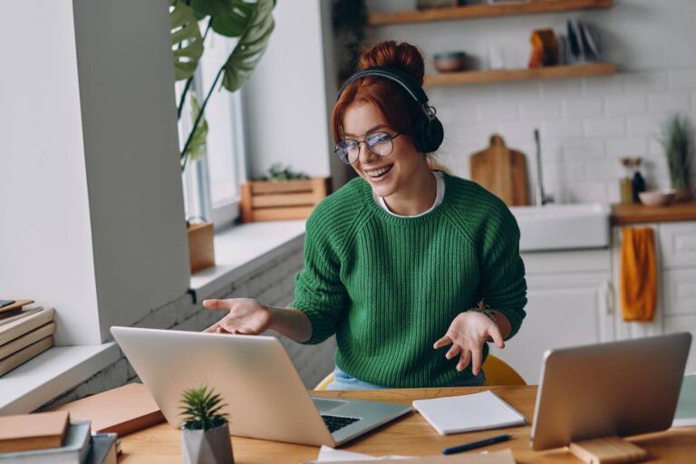 A young woman working from home in a green sweater at a bun. She's sitting at a desk with a laptop and a tablet.
