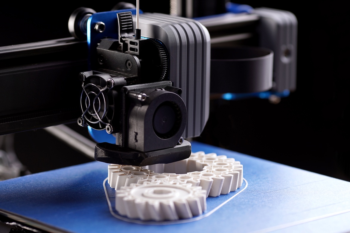 A black 3d printer generates large and small white plastic gears next to each other on top of a blue base.