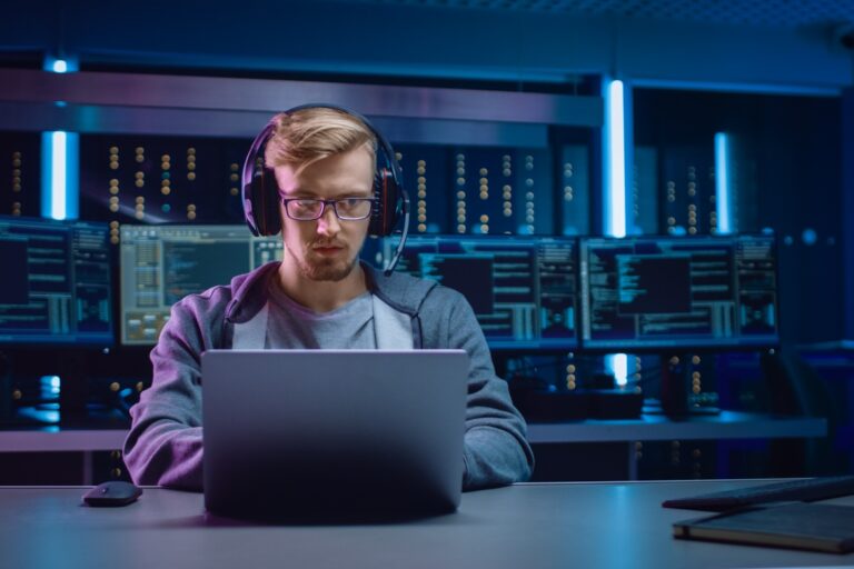 A man wearing glasses and a headset sits at a table and types on a laptop in a dark room with four large monitors behind him.