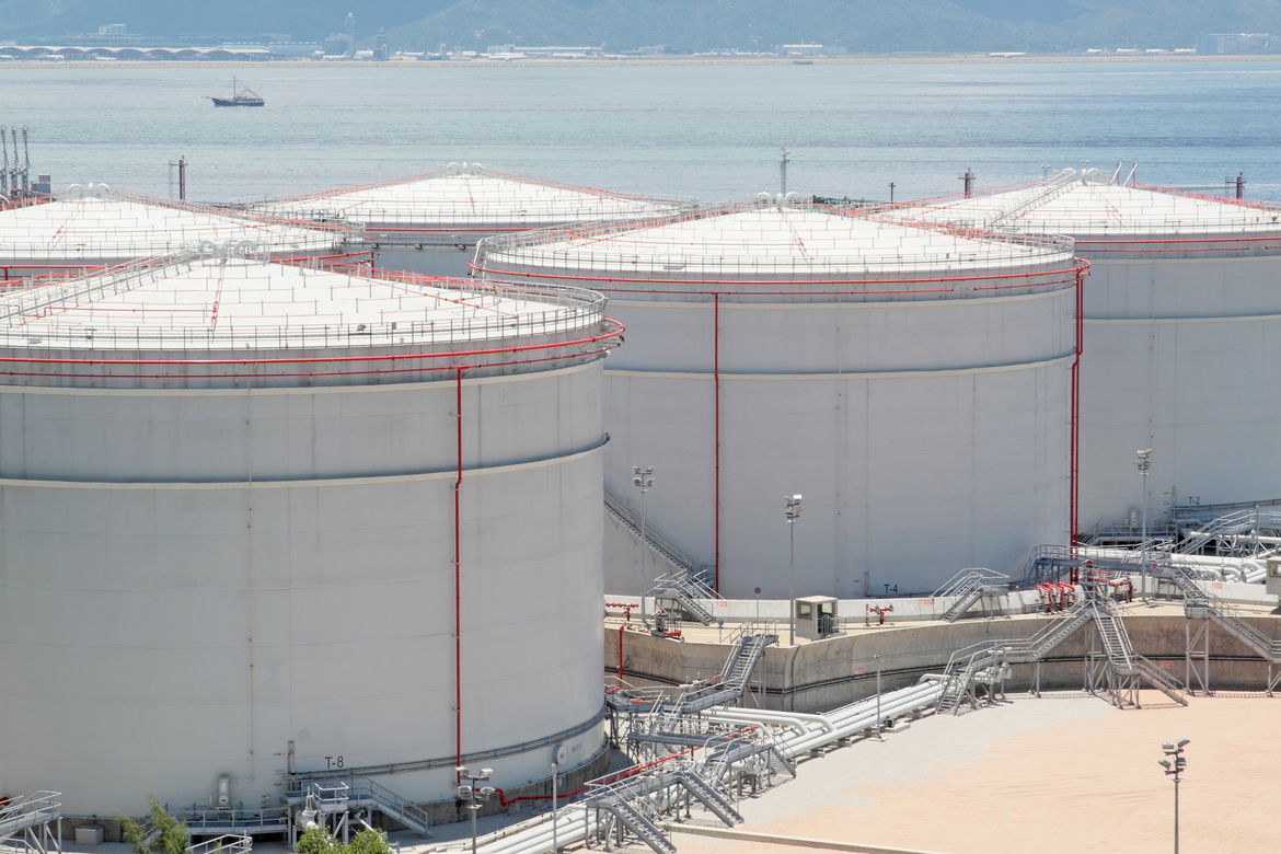 Large industrial tank in an empty plot of land near a large body of water in the background. There's metal bars on the tanks.