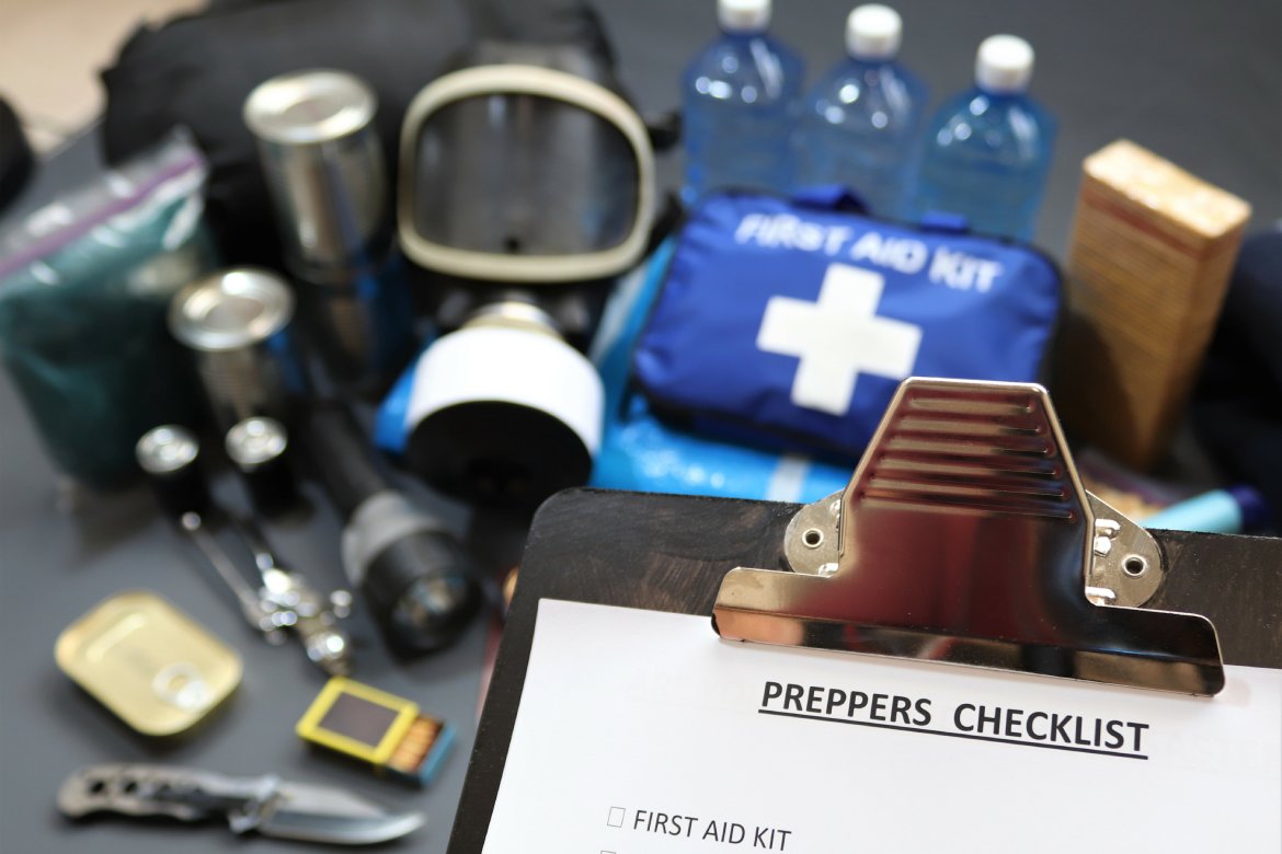 A clipboard labeled "preppers checklist" is held in the air above a table covered with various survival gear and supplies.