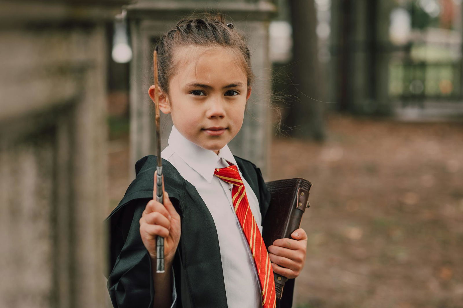 A young girl dressed as a wizard holding a wand and book in an outdoor setting, showcasing fantasy themed cosplay.