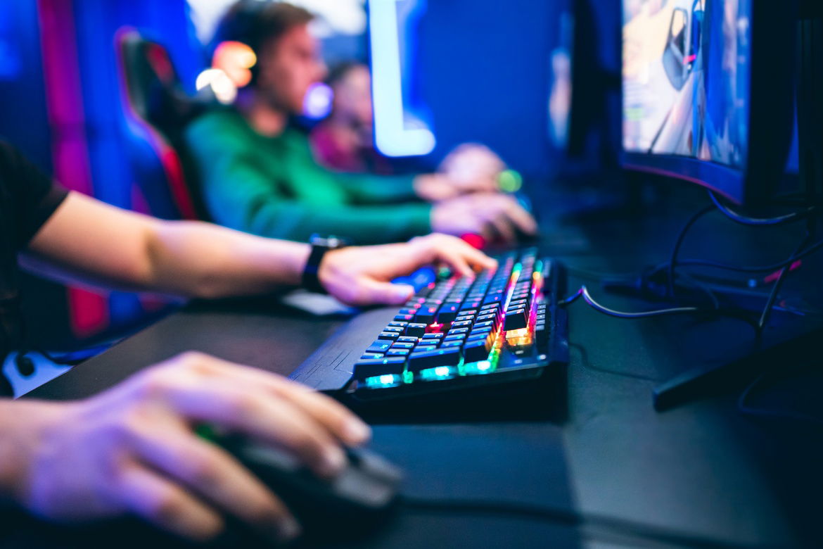 A group of video gamers sitting in a row while playing pc games in front of large monitors and colorful keyboards.