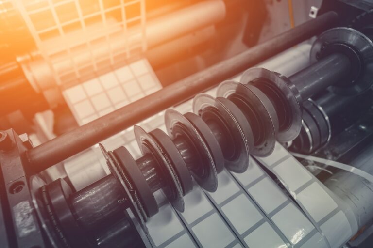 A rotary die cutting machine cuts long strips of square adhesive labels as they pass below the rotating blade.