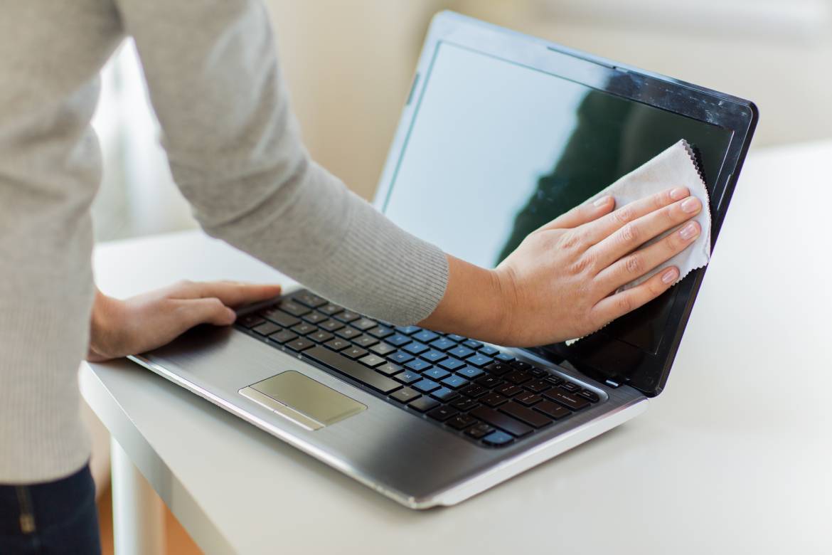 A close-up of someone's hands cleaning a laptop screen on their workstation using a disinfectant cloth.