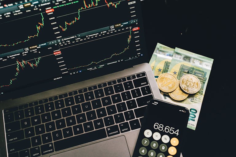 A laptop displaying stock charts with bitcoin, euros, and a cellphone calculator, showcasing financial analysis.