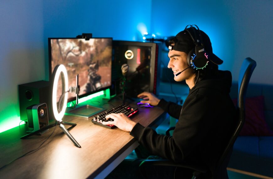 Young man wearing headset with microphone playing video game on computer with dual monitors and ring light.