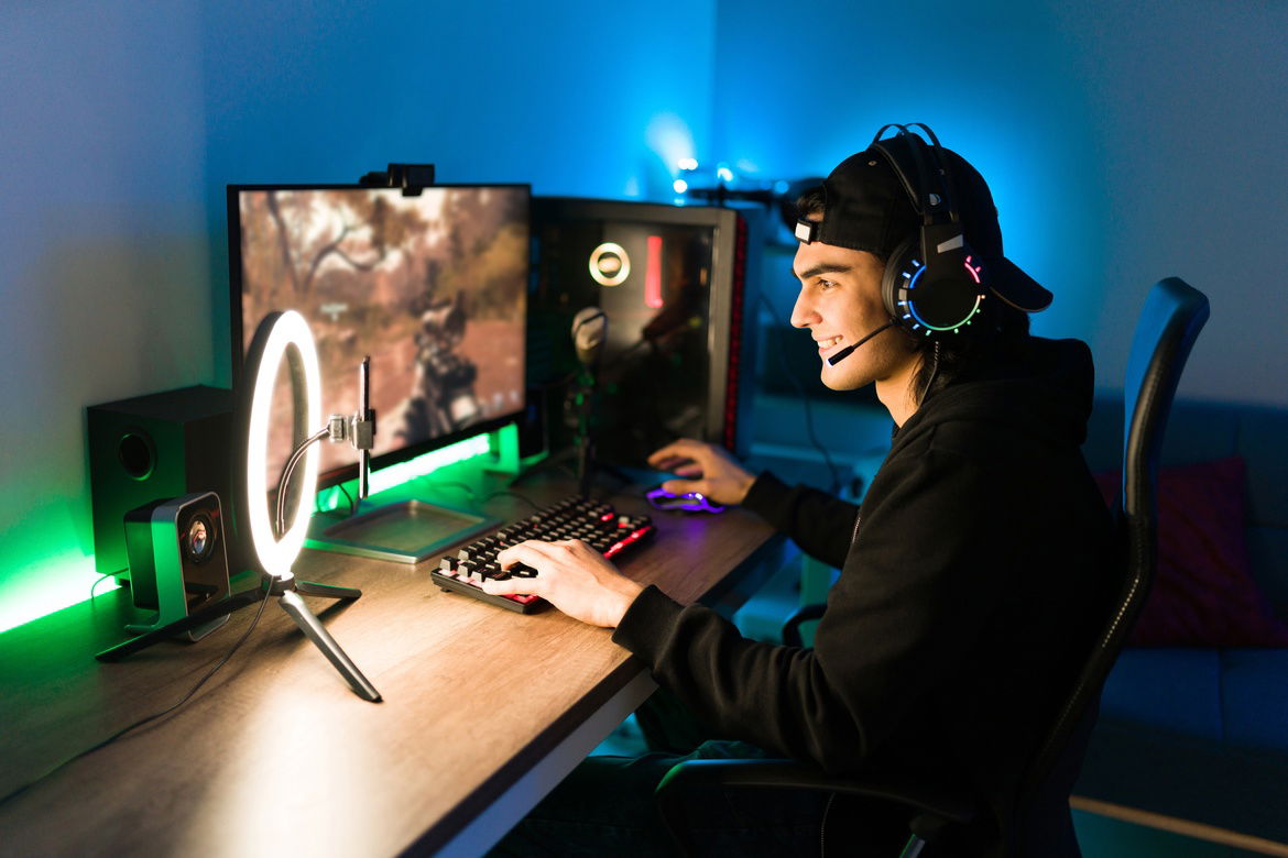 Young man wearing headset with microphone playing video game on computer with dual monitors and ring light.