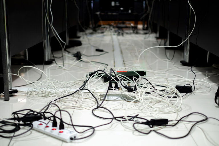 A long hallway full of tangled wires, cords, and power strips on the floor. Wires are hanging down from the walls.