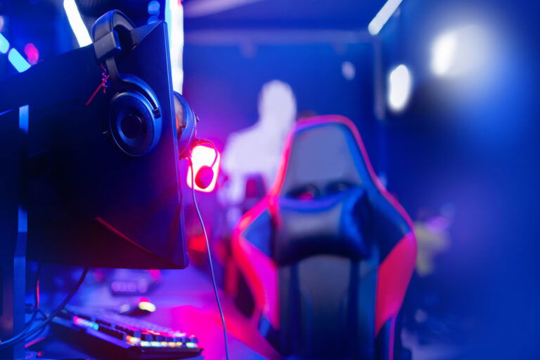 A gaming room features a red and black ergonomic chair, headphones, and colorful lights surrounding the space.