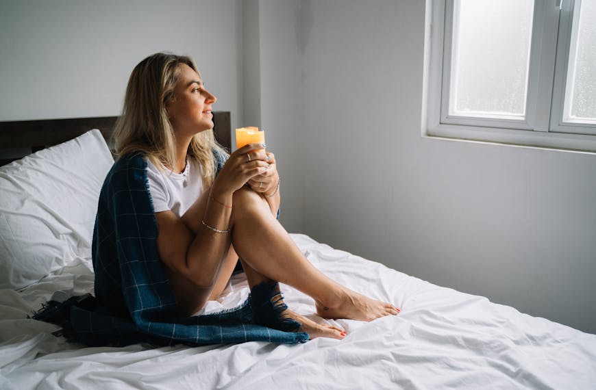 Woman sitting on bed with candle wrapped in a cozy blanket, enjoying morning tranquility.
