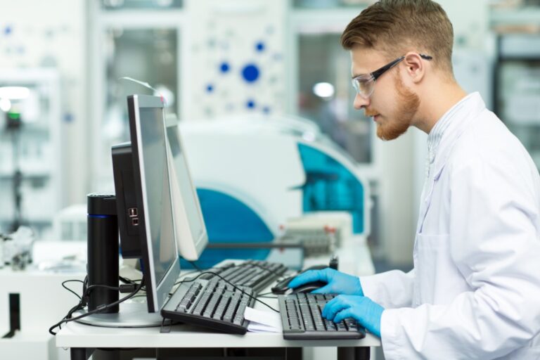 A young man wearing safety glasses, a white robe, and blue gloves, using a computer inside a lab environment.