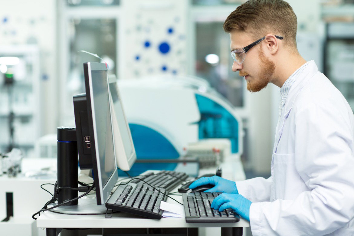A young man wearing safety glasses, a white robe, and blue gloves, using a computer inside a lab environment.