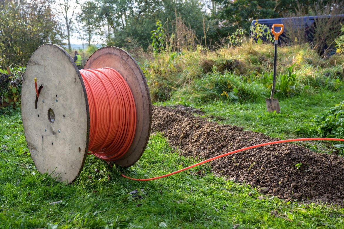 A spool of bright orange fiber wire sits in a grassy area off the road, ready for installation. There's a shovel next to it.