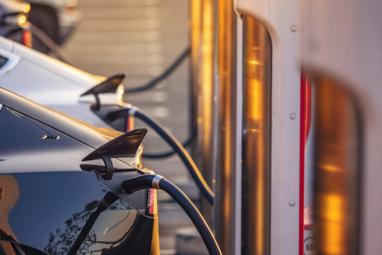 Two electric cars plugged into charging stations outdoors with warm sunlight reflecting on the vehicles and chargers.