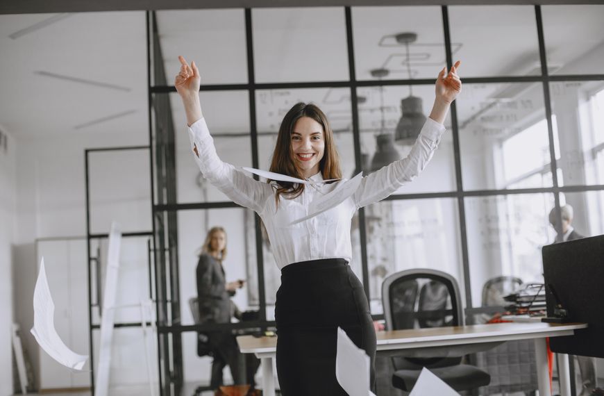 Smiling businesswoman in a modern office throws papers in the air, expressing joy and success.