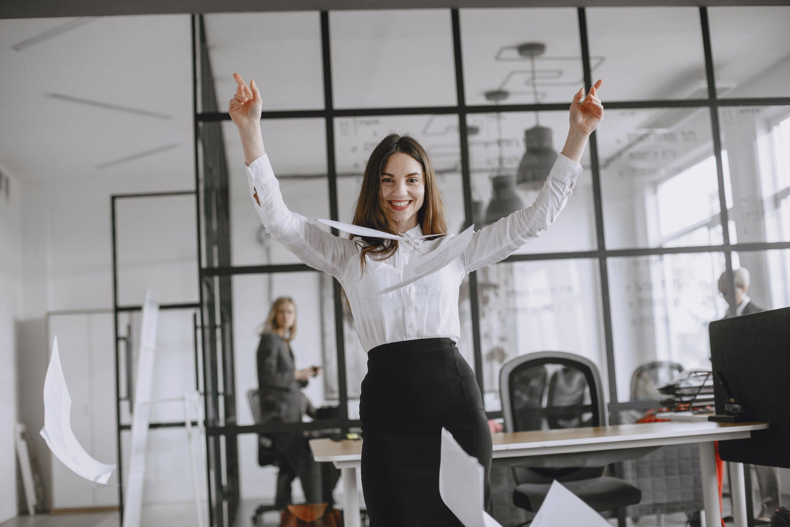 Smiling businesswoman in a modern office throws papers in the air, expressing joy and success.