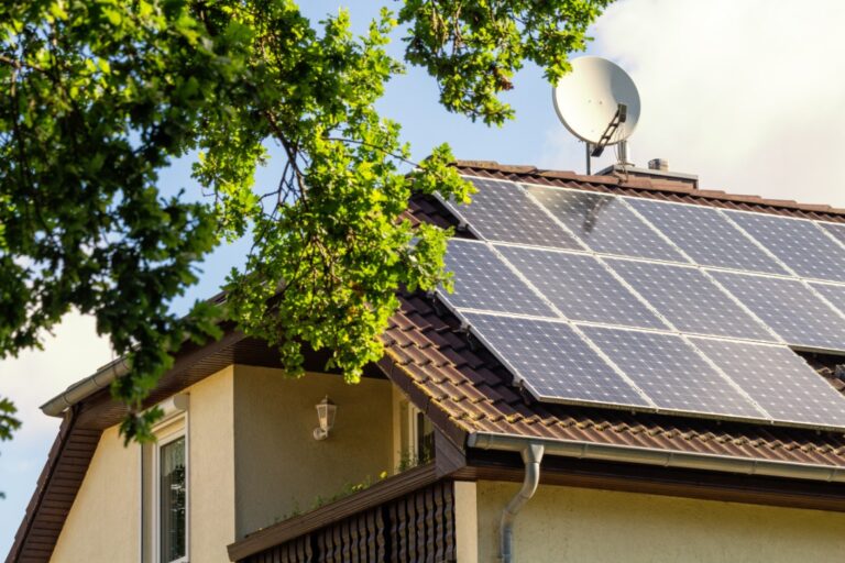 A two-story home with three long rows of solar panels on the roof, next to a dish antenna and a large tree.
