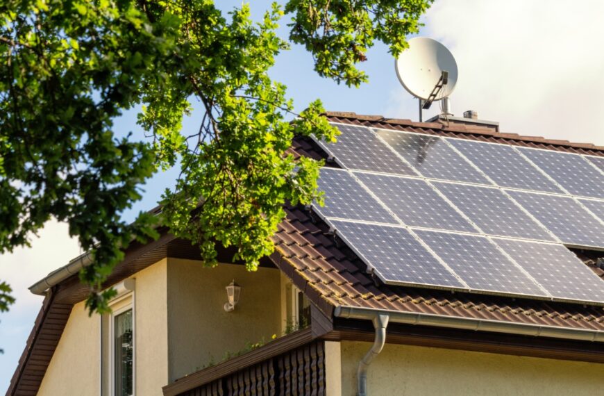 A two-story home with three long rows of solar panels on the roof, next to a dish antenna and a large tree.