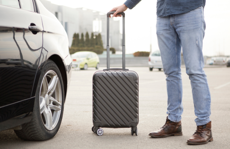 The back wheel of a car in an airport parking lot next to a man's legs as one hand rests on a rolling suitcase's handle