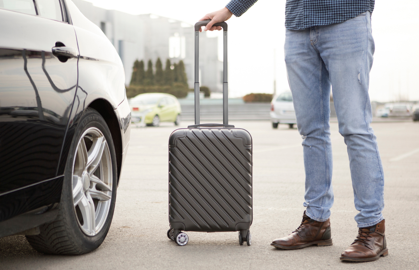 The back wheel of a car in an airport parking lot next to a man's legs as one hand rests on a rolling suitcase's handle