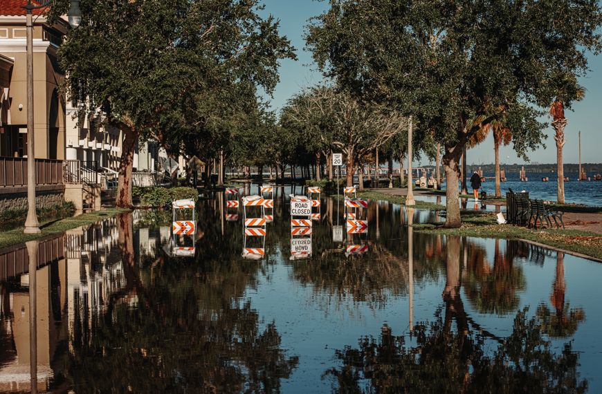 A flooded street with road closed signs reflecting in the water along a tree-lined sidewalk.