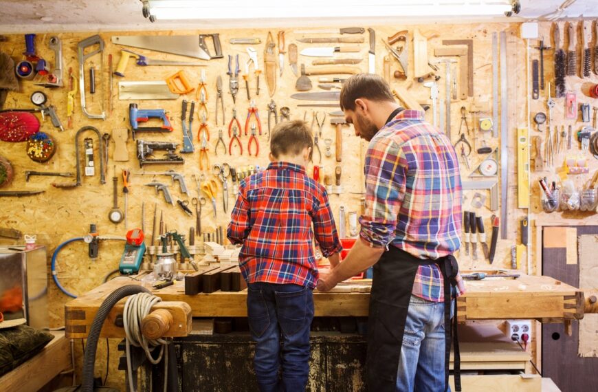 A father-son duo wear flannel shirts as they stand at their home workshop in the garage. The shelving has tools.
