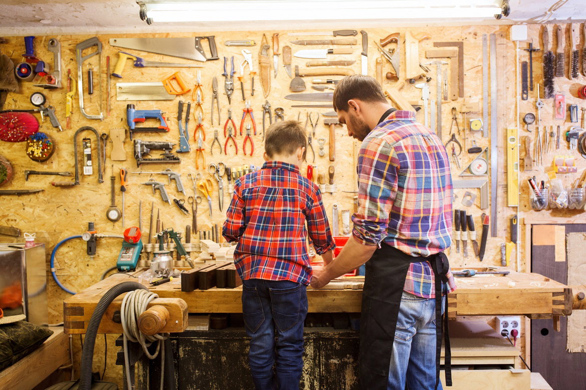 A father-son duo wear flannel shirts as they stand at their home workshop in the garage. The shelving has tools.
