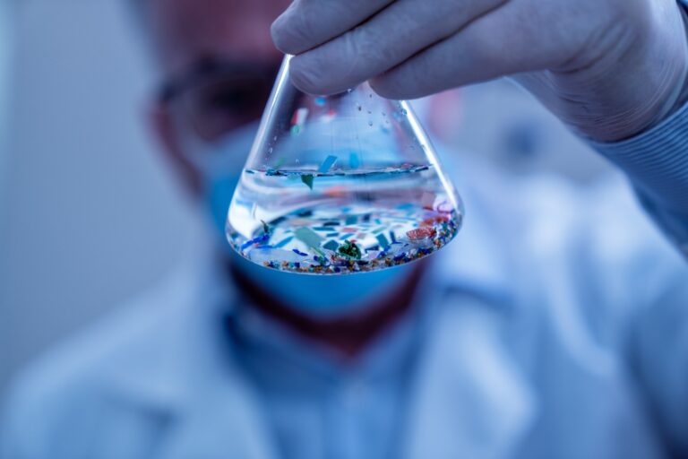 A close-up of a glass beaker held by a scientist in latex gloves and a lab coat. The beaker contains fluid and particles.
