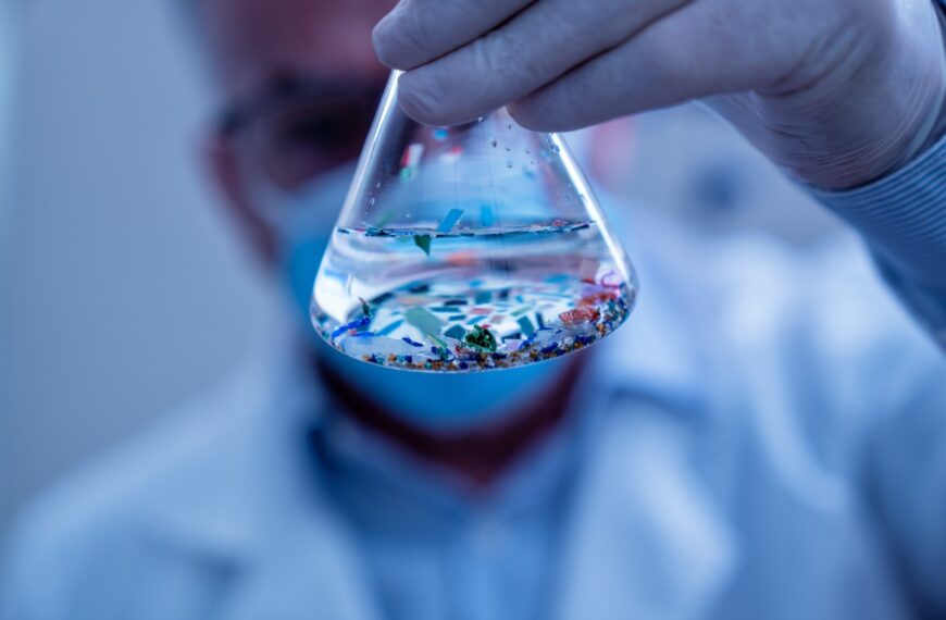A close-up of a glass beaker held by a scientist in latex gloves and a lab coat. The beaker contains fluid and particles.