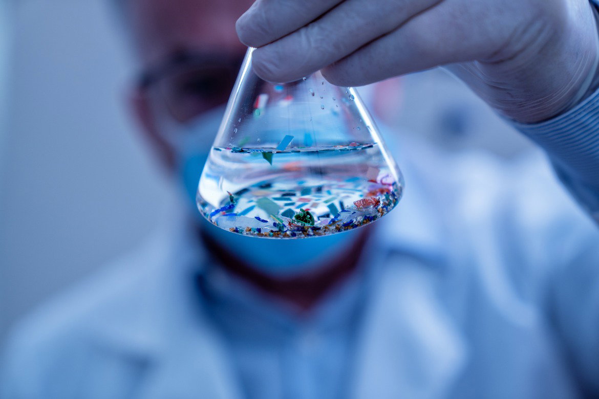 A close-up of a glass beaker held by a scientist in latex gloves and a lab coat. The beaker contains fluid and particles.