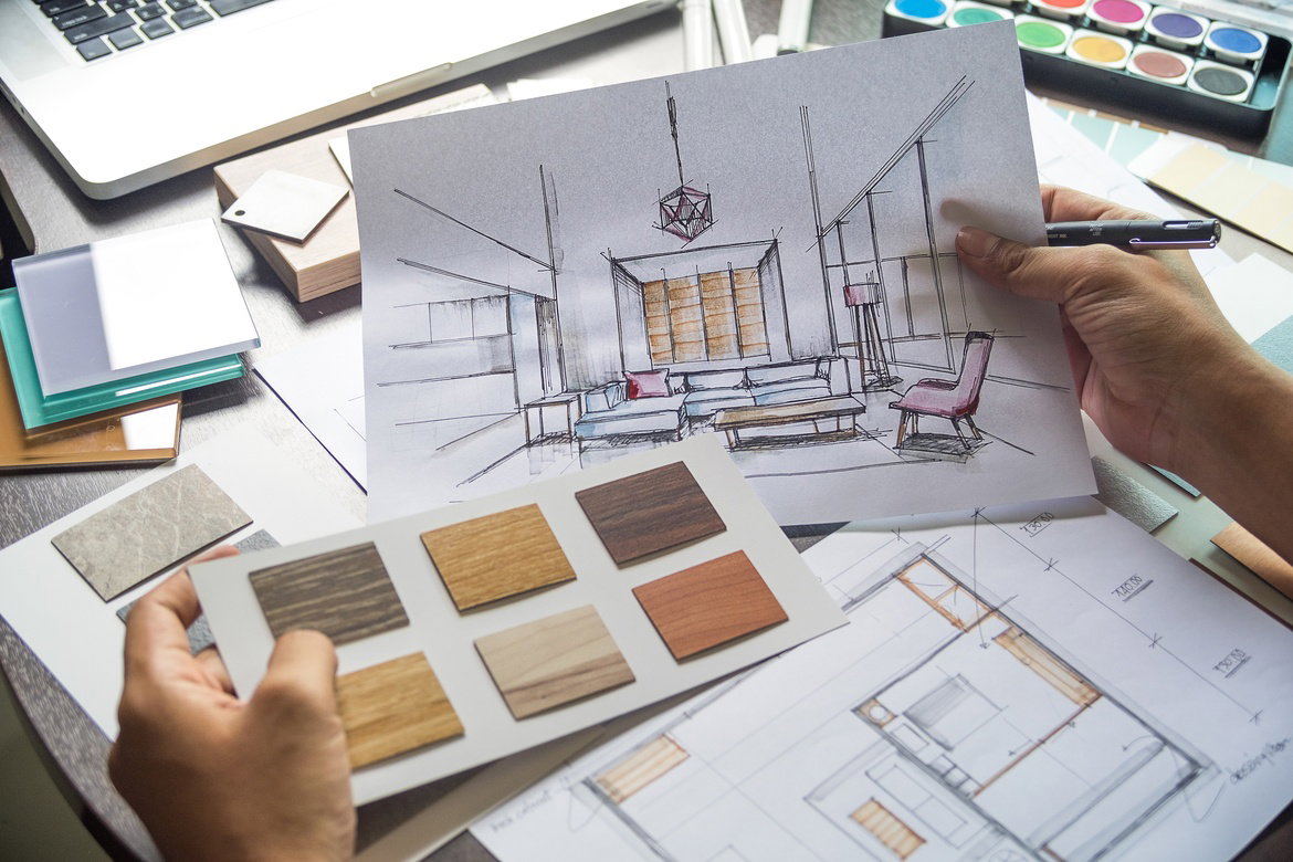 A person sits at a table full of blueprints and swatches. They hold samples and ideas in their hands.