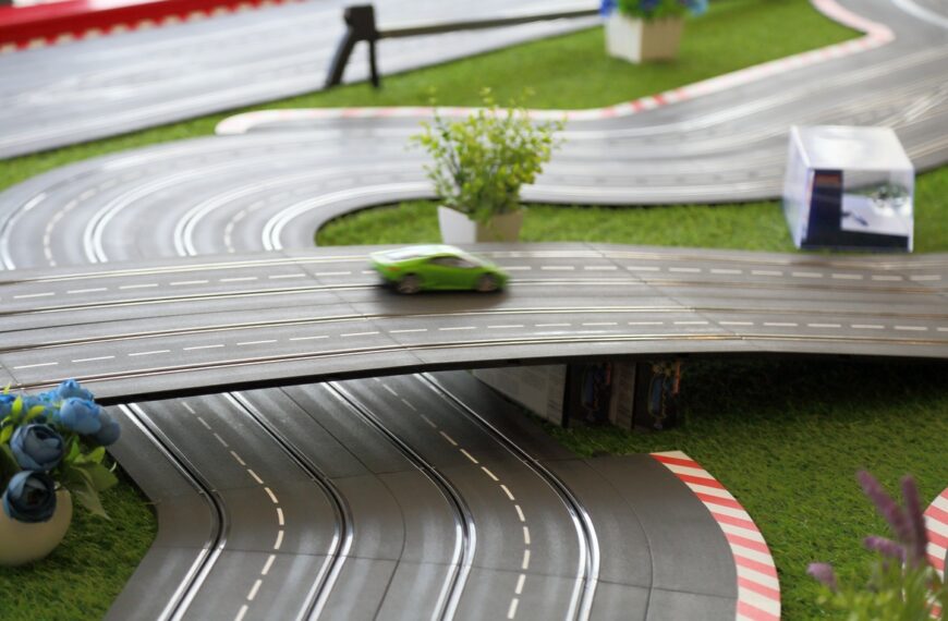 A green slot car racing around a multilevel track with curves, straightaways, landscaping, and an elevated bridge.